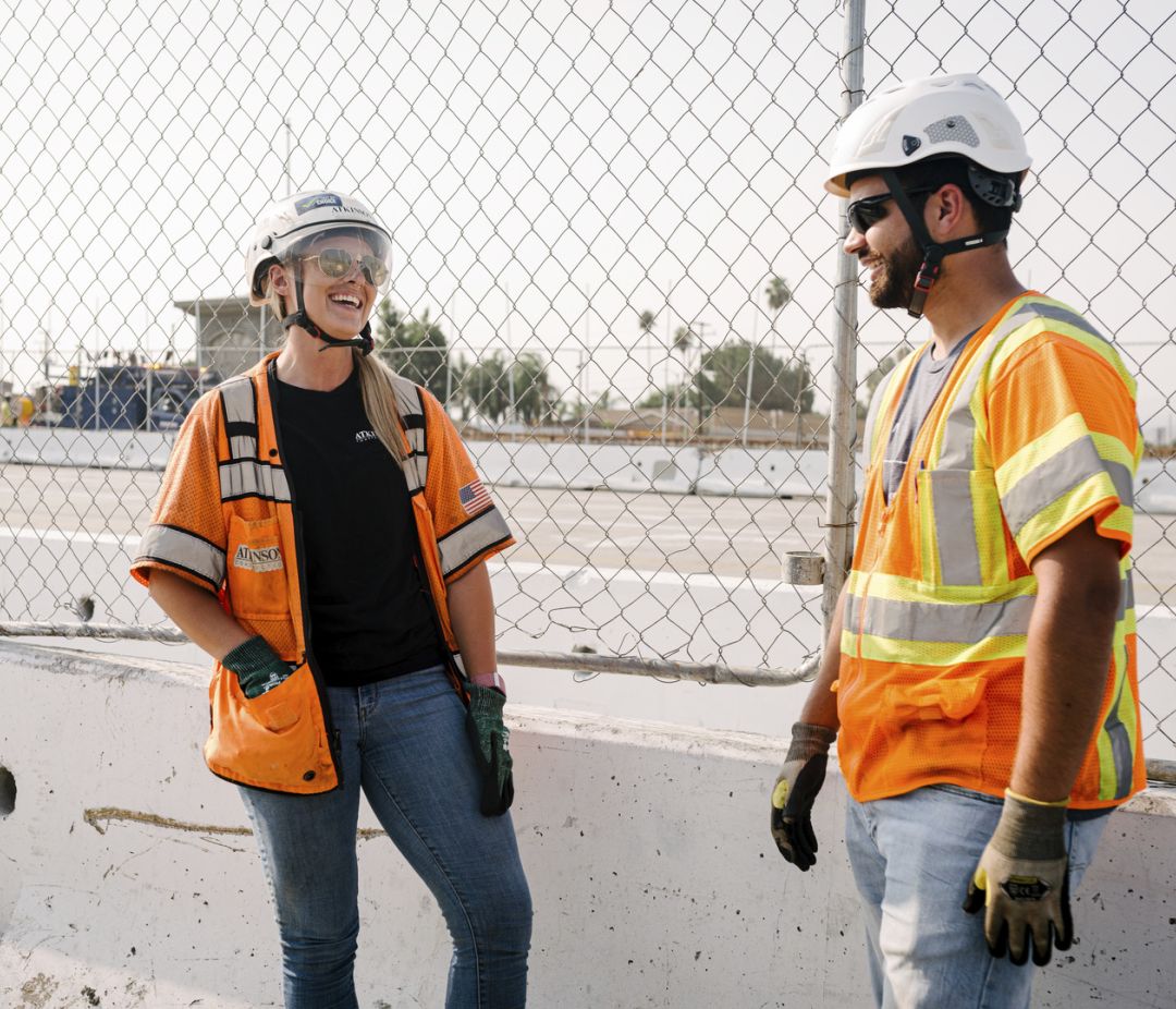 Two Atkinson team members, who are wearing full PPE, talk on a jobsite with metal fencing
